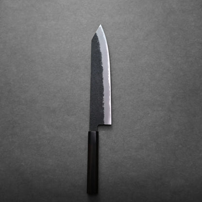 A kiritsuke Japanese chef's knife with a dark textured blade and handle stands vertically against a plain gray background.