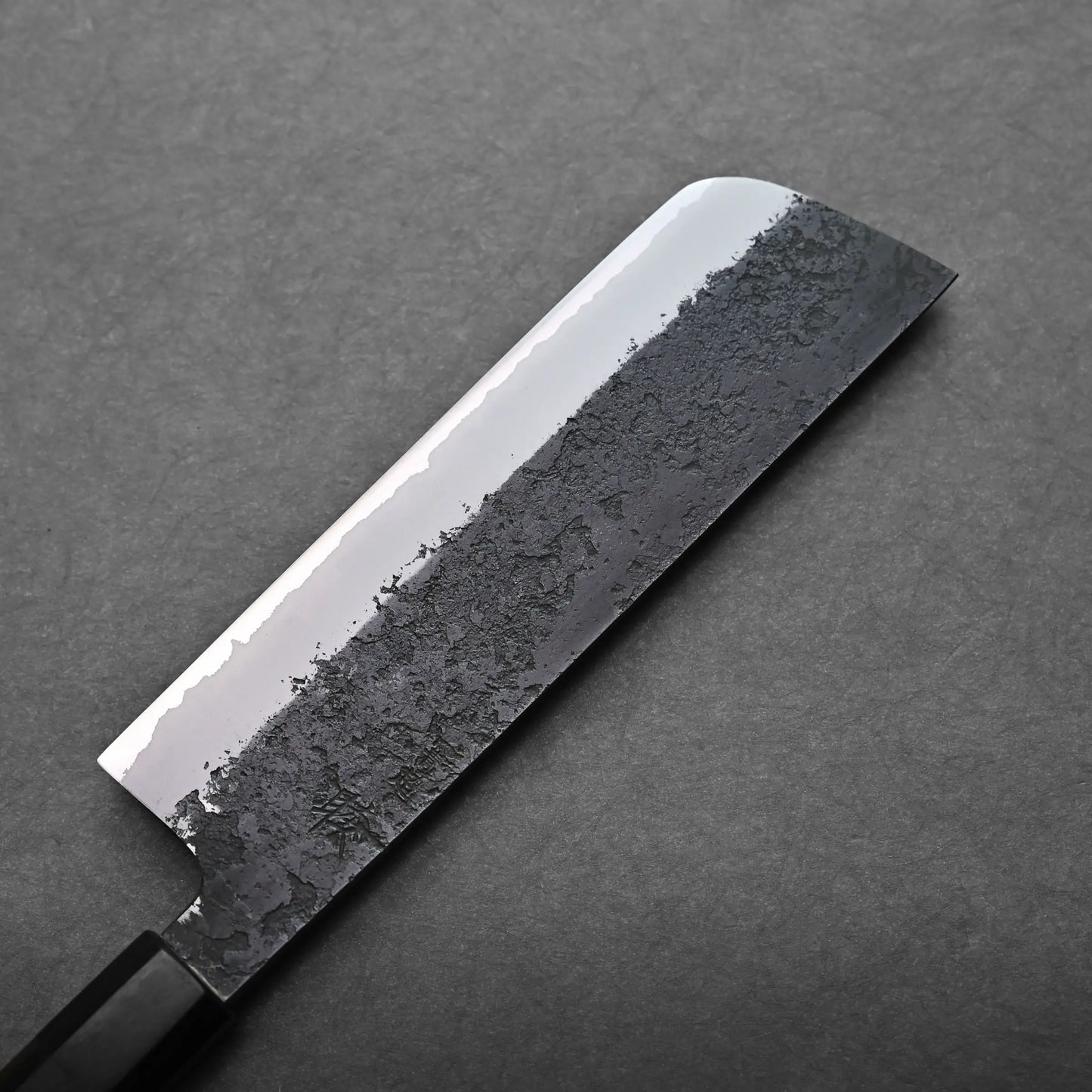 A close-up view of a Japanese vegetable knife with a rectangular, hammered blade resting on a dark gray surface.