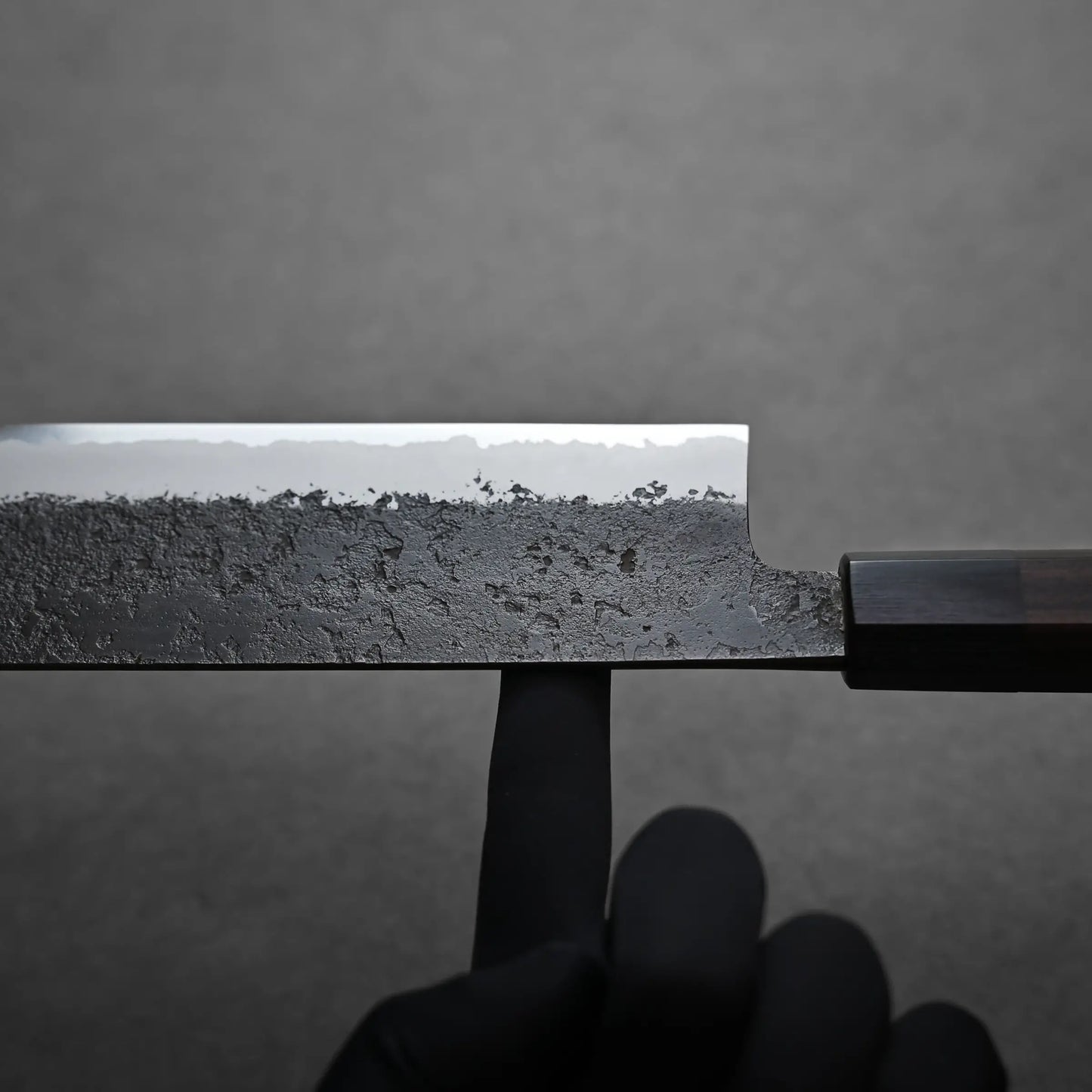 A gloved hand balances a nakiri knife by its blade on an index finger. The knife features a textured, rustic-looking blade and a dark wooden handle, set against a plain gray background. It shows the balance point.