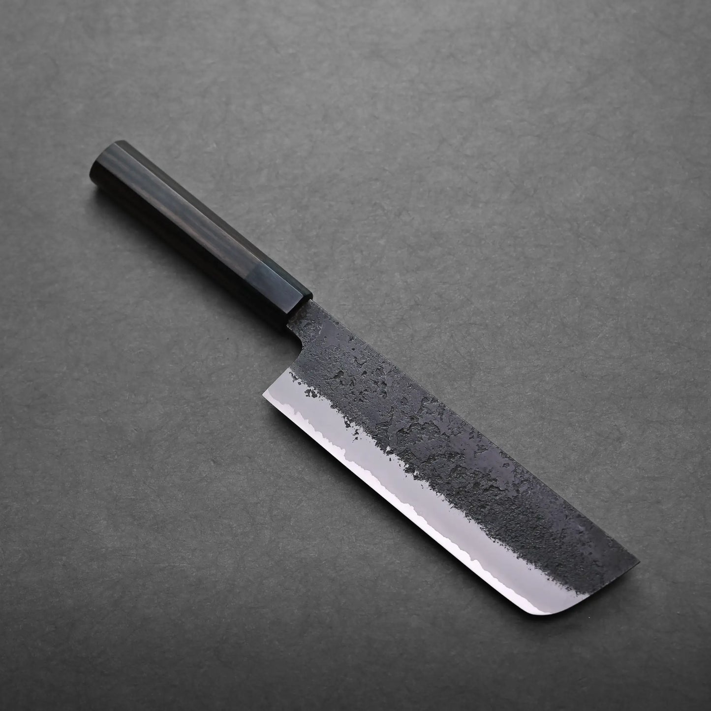 A Japanese vegetable knife with a black handle and a dark, textured blade lies on a gray surface. The nakiri knife features a straight edge and a shiny, polished lower section.