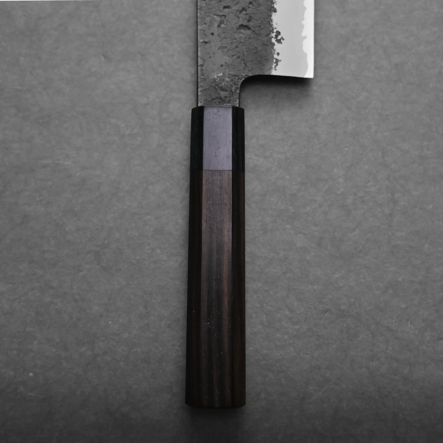 Close-up of the handle and lower blade of a nakiri knife with a dark wooden handle and a textured, partially polished blade, set against a plain gray surface.