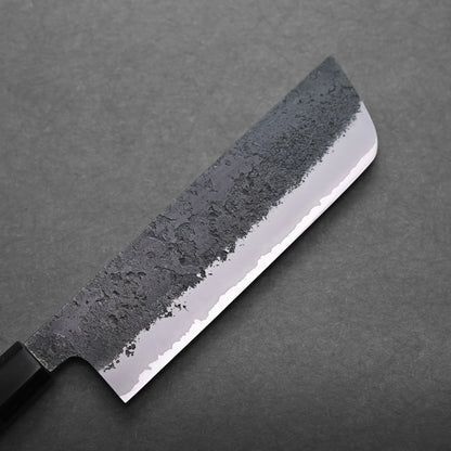 A close-up view of a Japanese vegetable knife with a dark, textured blade and a polished, sharp edge, resting on a plain grey surface.