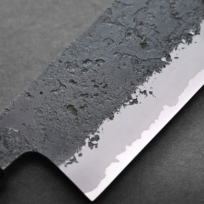 Close-up of a Japanese vegetable knife blade showing a sharp edge with a textured, hammered black finish on the upper part. This nakiri knife rests on a dark, smooth surface.