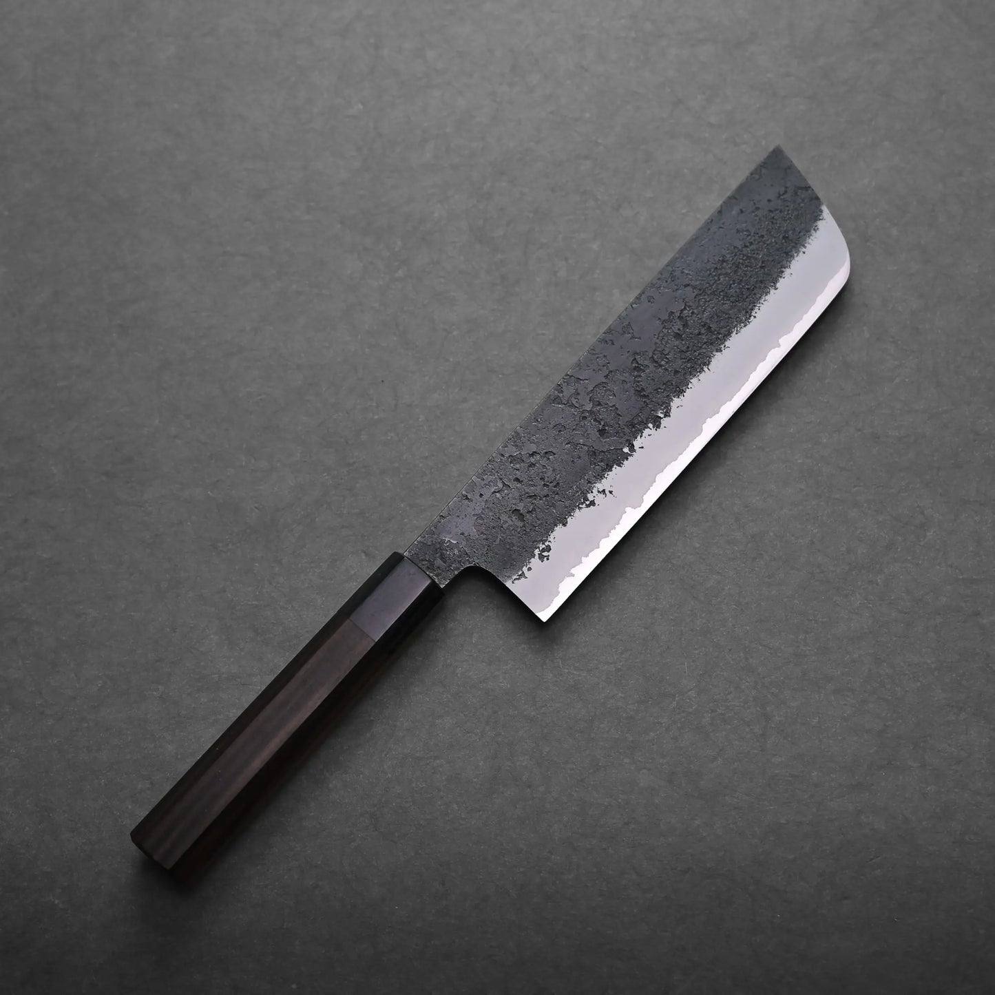 A Japanese vegetable knife with a dark wooden handle and a rectangular, partially textured blade lies on a dark, flat surface.