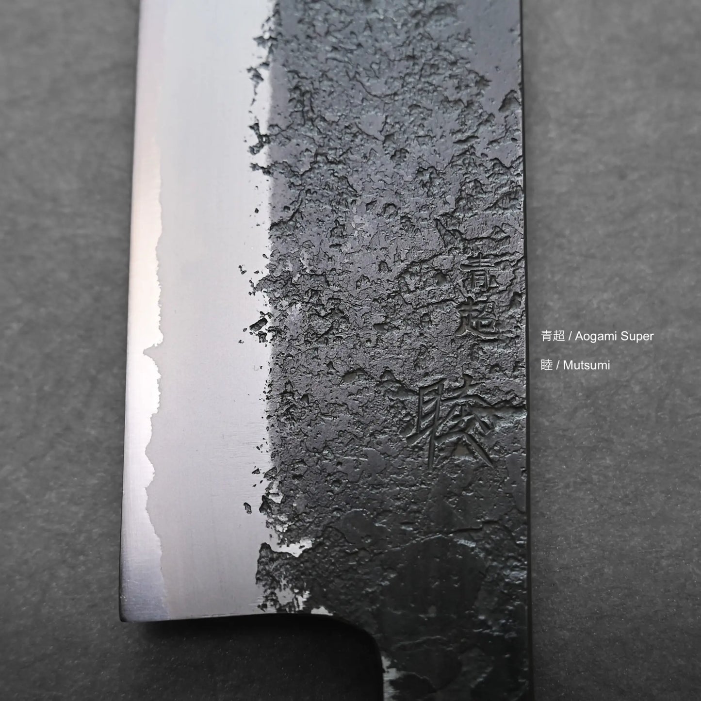 Close-up of a nakiri knife blade with a textured, mottled upper half and smooth, polished edge below. Japanese characters and the words "Aogami Super" and "Mutsumi" are engraved on the Japanese vegetable knife.