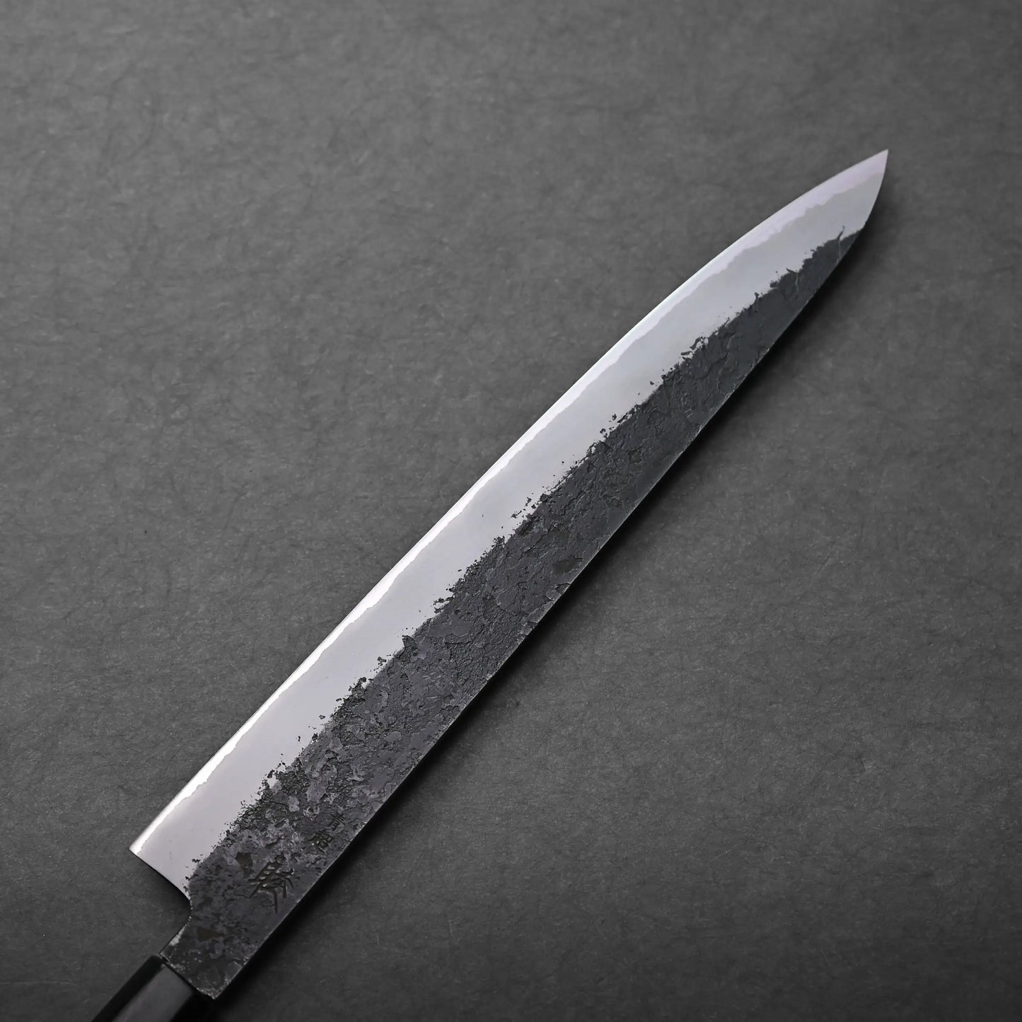 A close-up of a Japanese slicing knife with a single bevel edge and a dark, textured finish along the blade, placed on a dark gray surface.