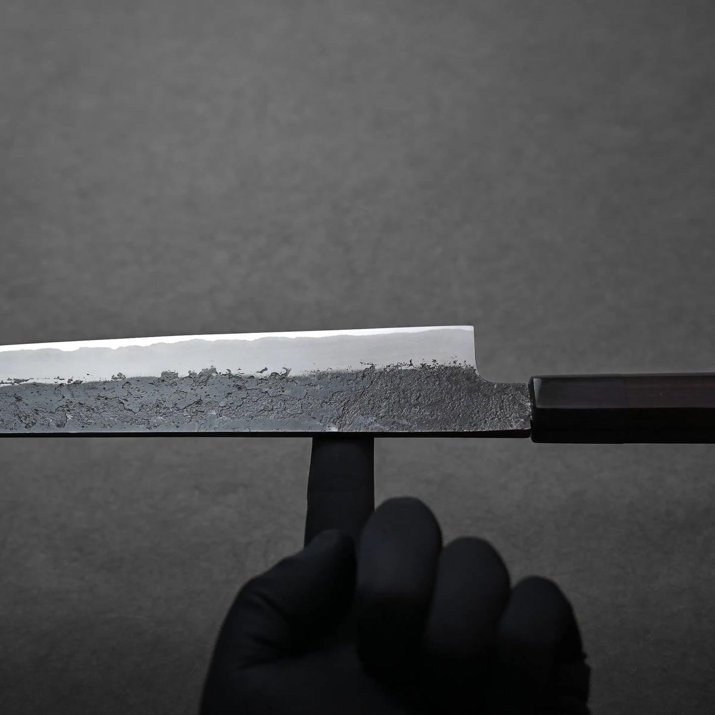 A hand in a black glove balances a large Japanese sujihiki knife on one finger, its blade facing upwards against a plain dark background. It shows the balance point.