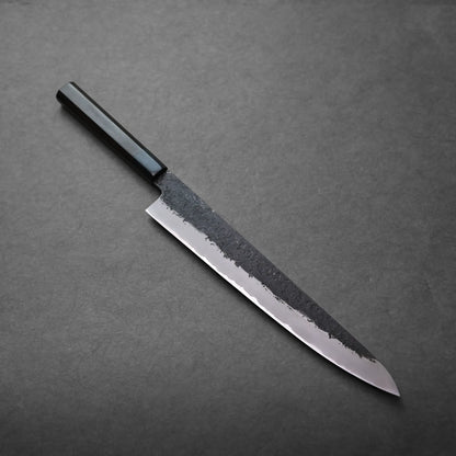 A Japanese slicing knife with a dark handle and polished blade lies on a flat, dark gray surface. The sujihiki knife features a textured, blackened upper blade and a razor-sharp edge.