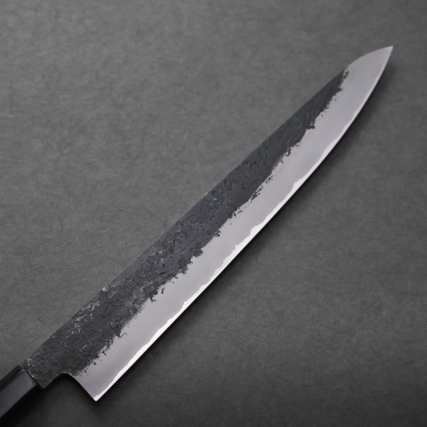 A close-up of a Japanese slicing knife with a long, narrow blade, featuring a textured, dark upper edge and a sharp, polished cutting edge, placed on a dark gray surface.