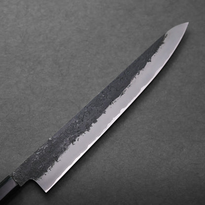 A close-up of a Japanese slicing knife with a long, narrow blade, featuring a textured, dark upper edge and a sharp, polished cutting edge, placed on a dark gray surface.