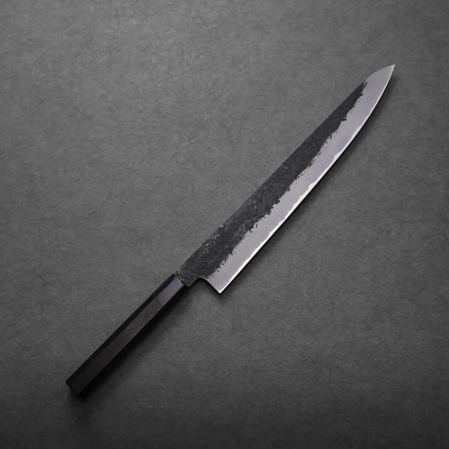 A Mutsumi Hinoura kurouchi nashiji AS sujihiki 270mm with a long, narrow blade and black handle rests on a dark grey textured surface. The blade features a raw, unfinished upper section and a polished cutting edge.