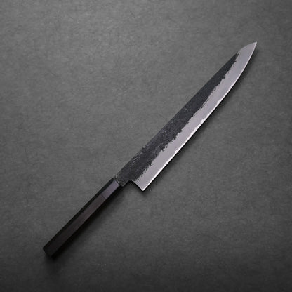 A Mutsumi Hinoura kurouchi nashiji AS sujihiki 270mm with a long, narrow blade and black handle rests on a dark grey textured surface. The blade features a raw, unfinished upper section and a polished cutting edge.