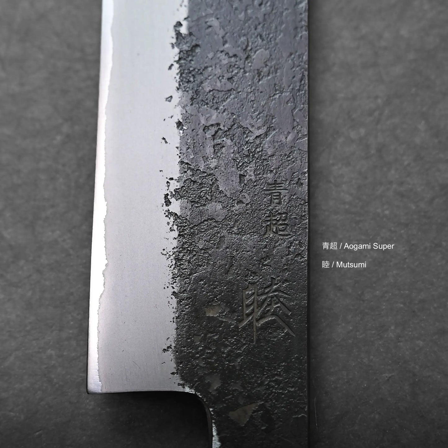 Close-up of a Japanese slicing knife blade with visible texture differences. The blade features engraved kanji characters and the words Aogami Super and Mutsumi, set against a dark background.