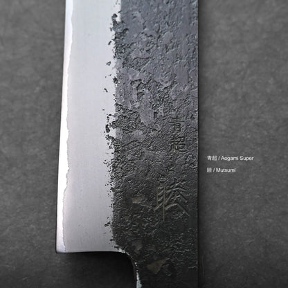 Close-up of a Japanese slicing knife blade with visible texture differences. The blade features engraved kanji characters and the words Aogami Super and Mutsumi, set against a dark background.