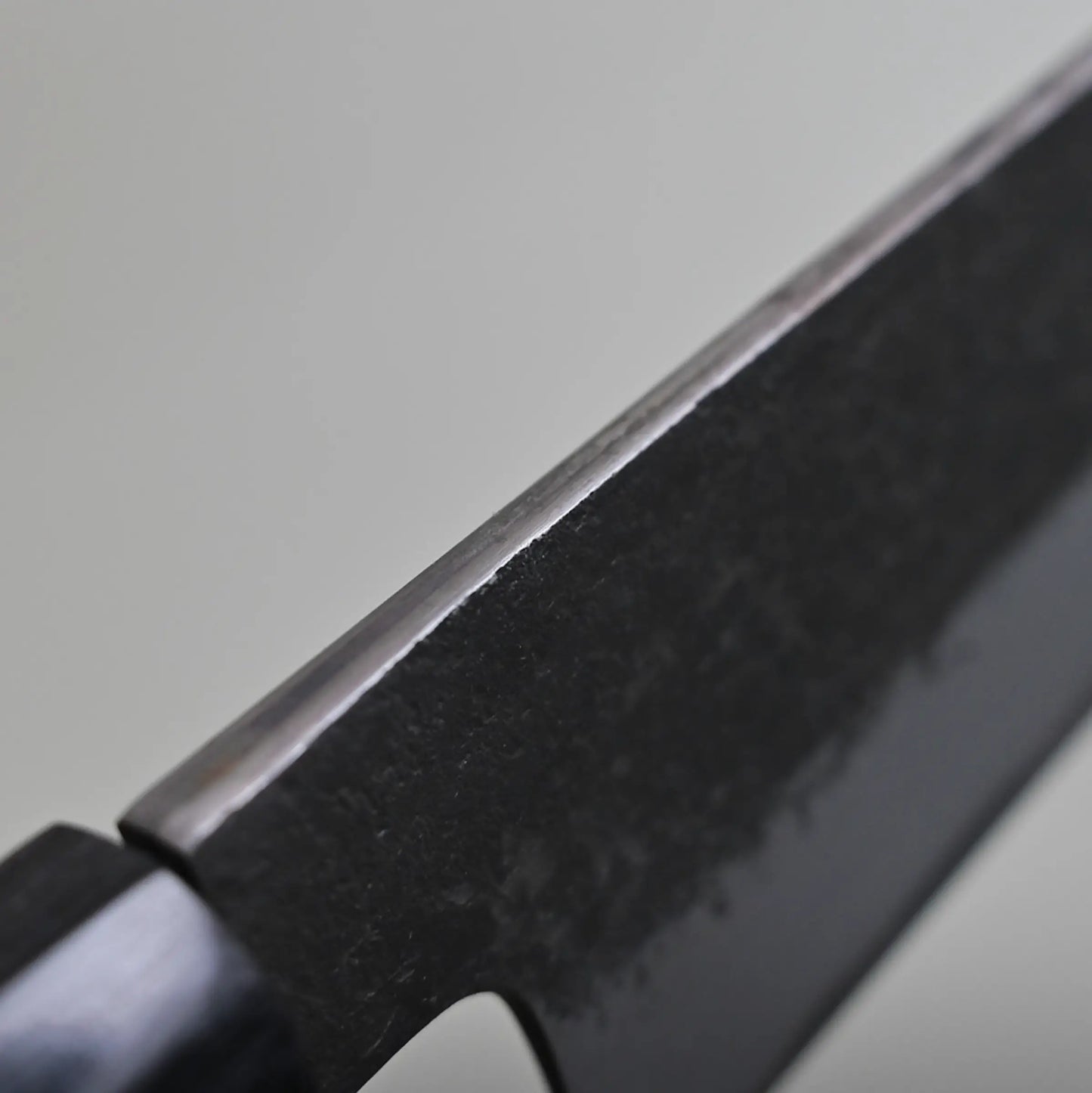 Close-up view of the spine finish of a dark-colored sujihiki knife, showing the edge and part of the handle against a plain light background.