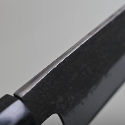 Close-up view of the spine finish of a dark-colored sujihiki knife, showing the edge and part of the handle against a plain light background.