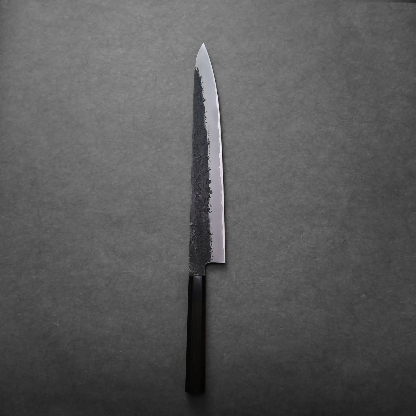 A single Japanese slicing knife with a dark, rustic blade and a black handle rests on a plain, dark gray surface.