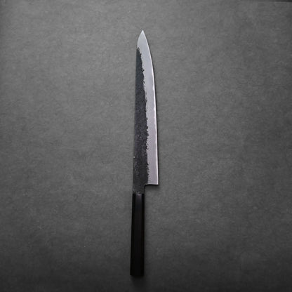 A single Japanese slicing knife with a dark, rustic blade and a black handle rests on a plain, dark gray surface.