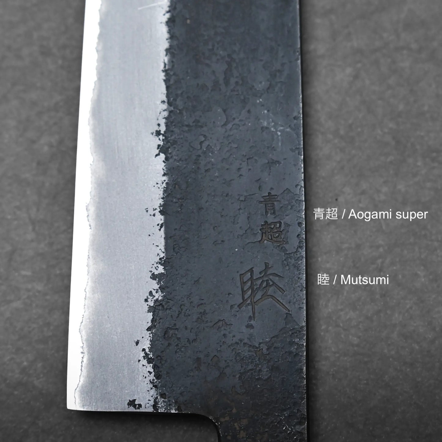 Close-up of a Hinoura gyuto blade highlighting the contrast between polished and textured steel, with Japanese characters engraved alongside product information text reading Aogami super and Mutsumi to the right.