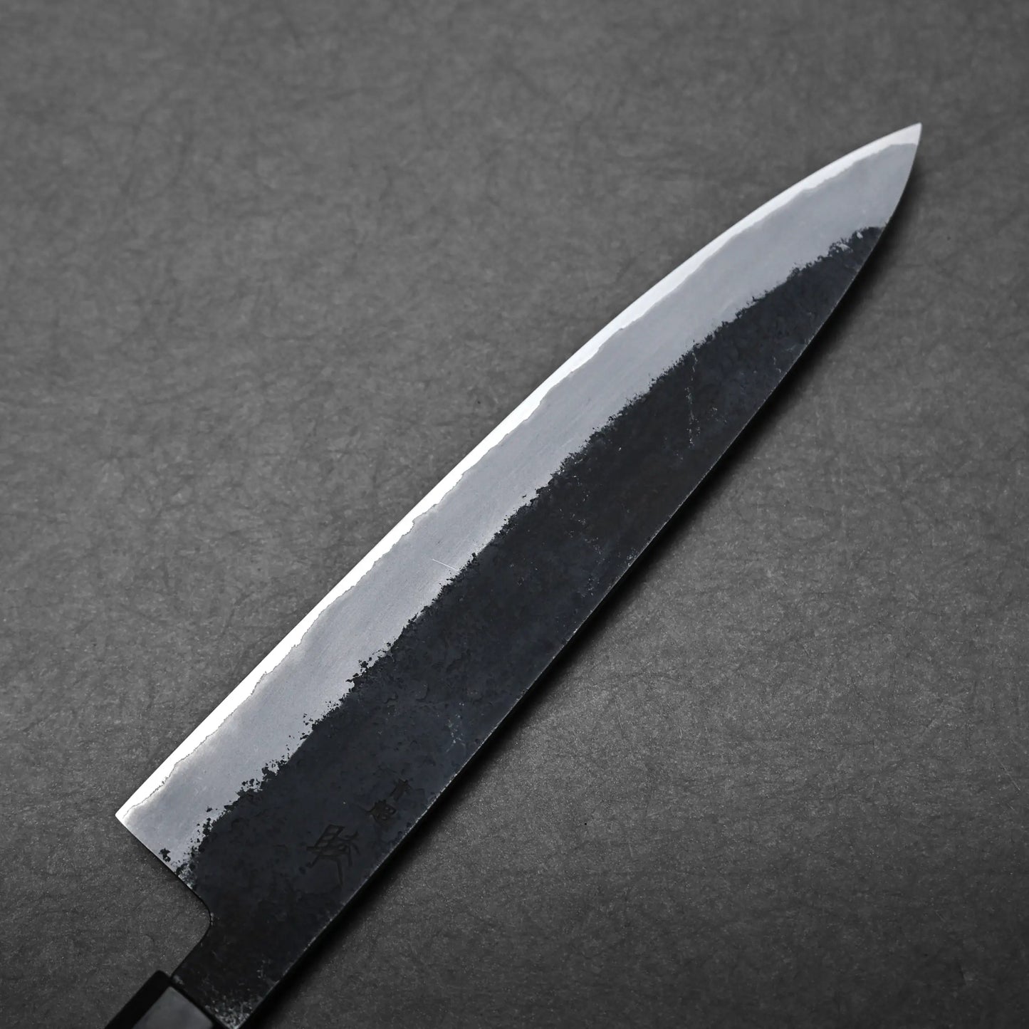 A close-up view of a Japanese chef's knife, featuring a dark, textured blade and polished finish, placed on a dark, textured surface.