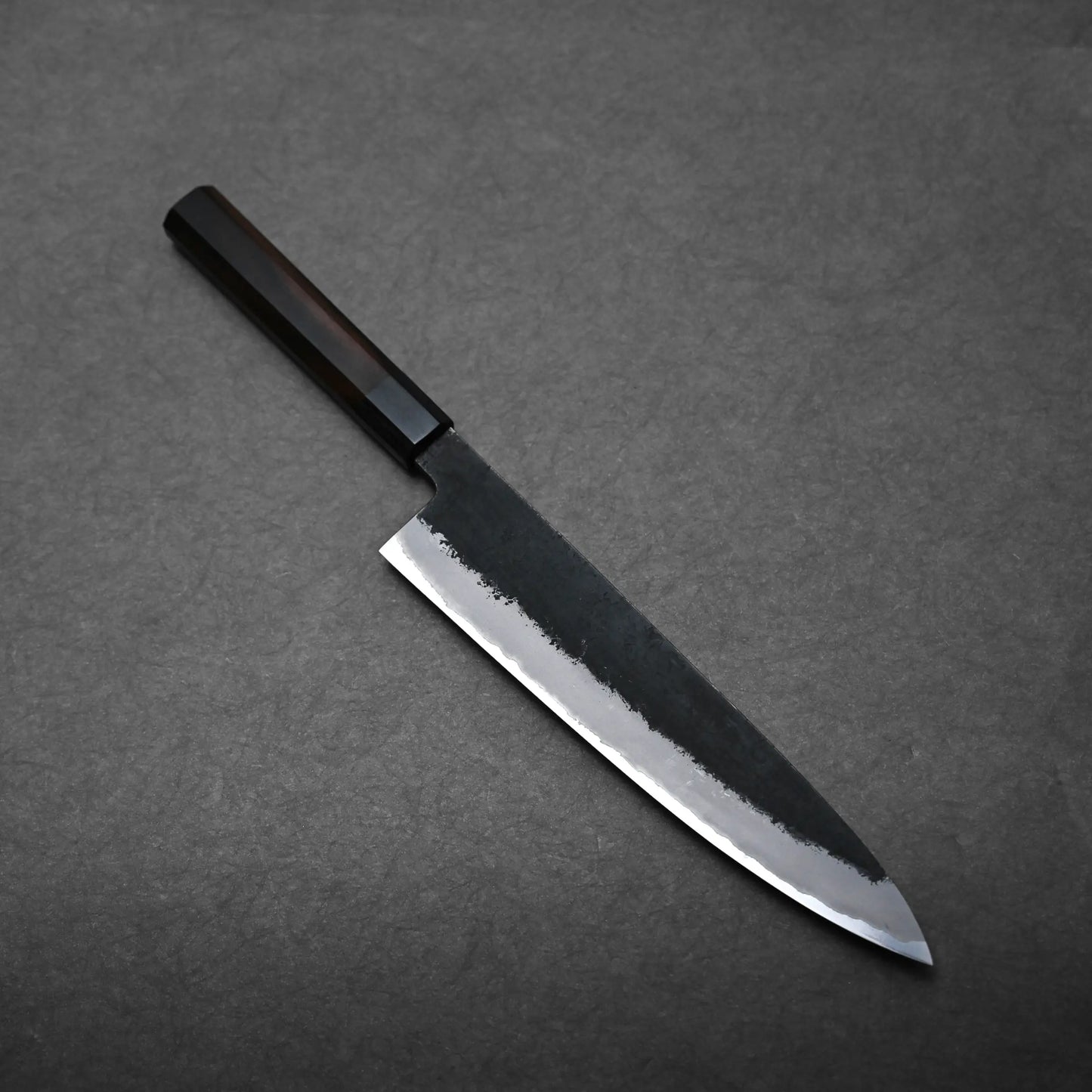 A Hinoura gyuto Japanese chef's knife with a dark handle and polished blade rests on a textured gray surface.