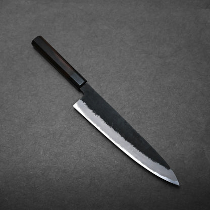 A Hinoura gyuto Japanese chef's knife with a dark handle and polished blade rests on a textured gray surface.