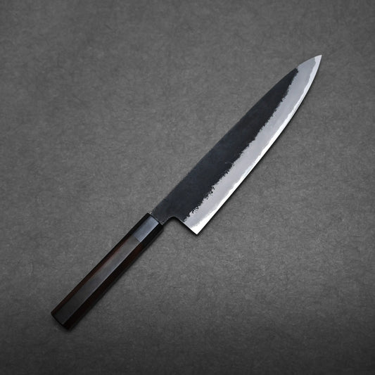 A Hinoura kurouchi nashiji AS (stainless clad) gyuto 240mm with a dark wood handle and a shiny, partially polished blade rests on a textured gray surface.