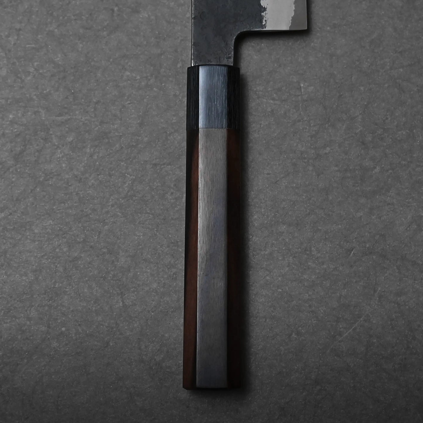Close-up of a dark wooden handle on a Hinoura gyuto, featuring a polished, faceted surface against a textured gray background. The blade of this Japanese chef's knife is partially visible at the top of the image.