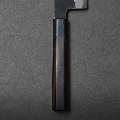 Close-up of a dark wooden handle on a Hinoura gyuto, featuring a polished, faceted surface against a textured gray background. The blade of this Japanese chef's knife is partially visible at the top of the image.