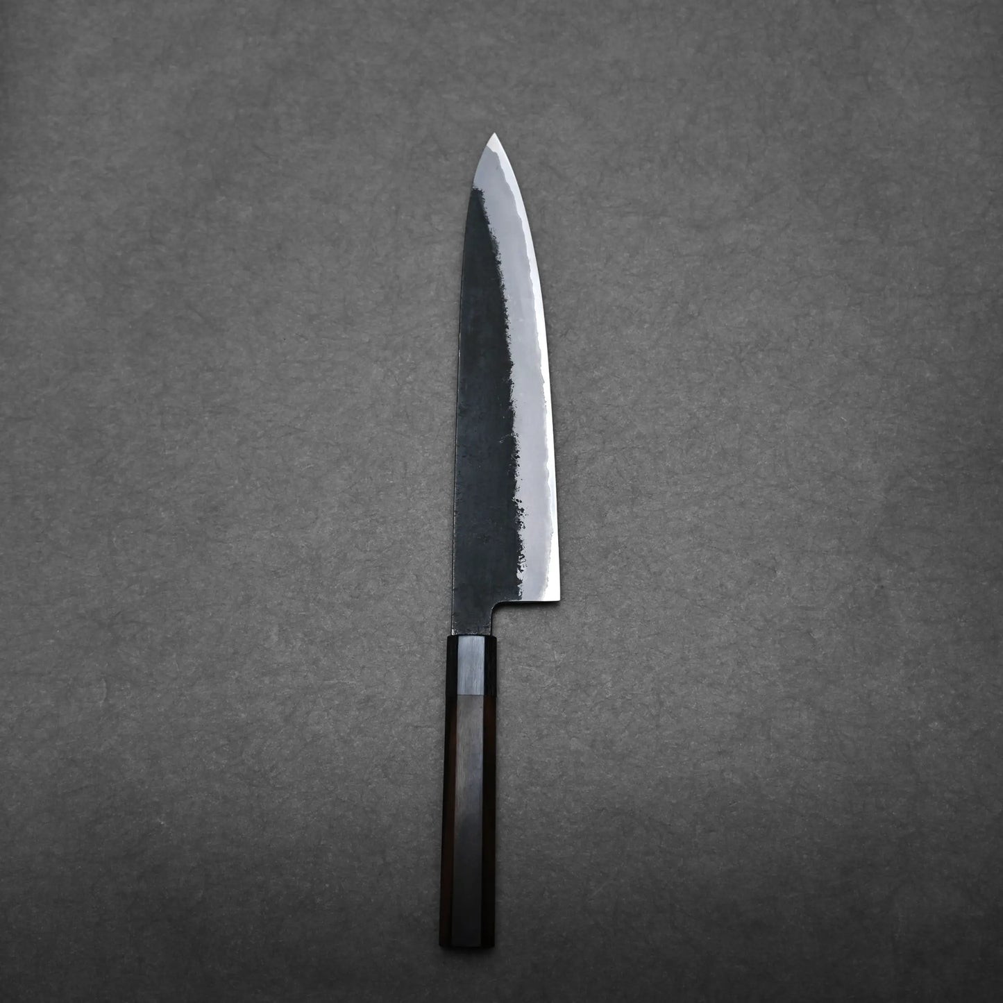 A single Hinoura gyuto Japanese chef’s knife with a dark handle and a partially polished, partially matte blade lies on a plain dark gray surface.