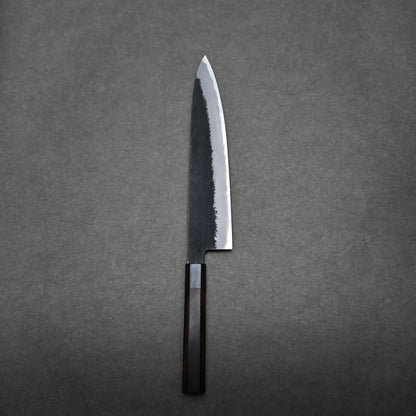 A single Hinoura gyuto Japanese chef’s knife with a dark handle and a partially polished, partially matte blade lies on a plain dark gray surface.
