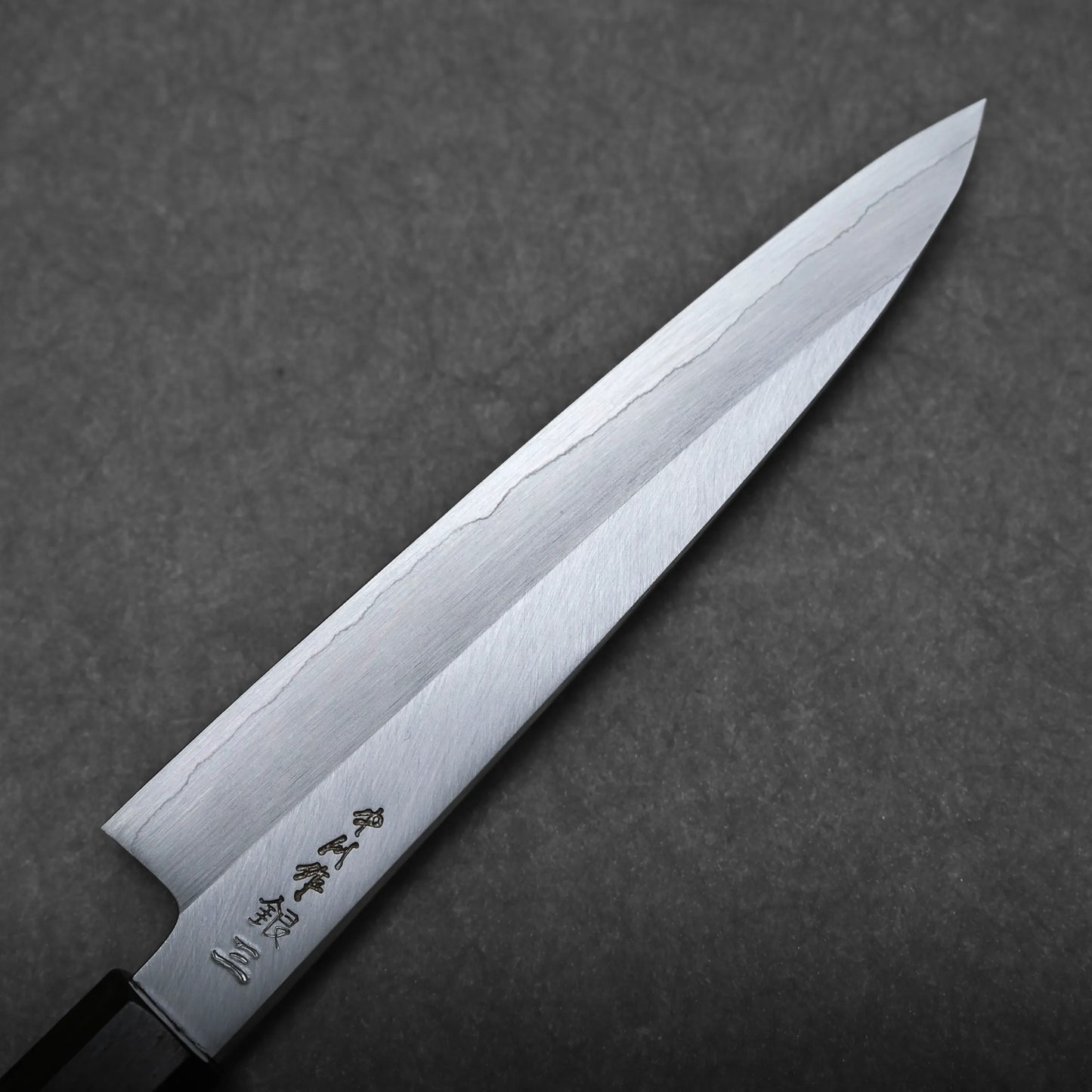 A close-up of a Japanese utility knife with a sharp, polished blade featuring subtle engraving and a visible hamon line, set against a gray textured background.