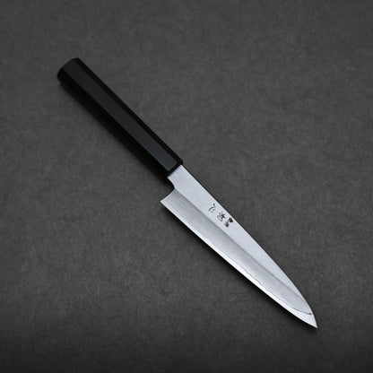 A sharp Japanese utility knife with a black handle and a polished stainless steel blade is laid diagonally on a dark, textured surface.