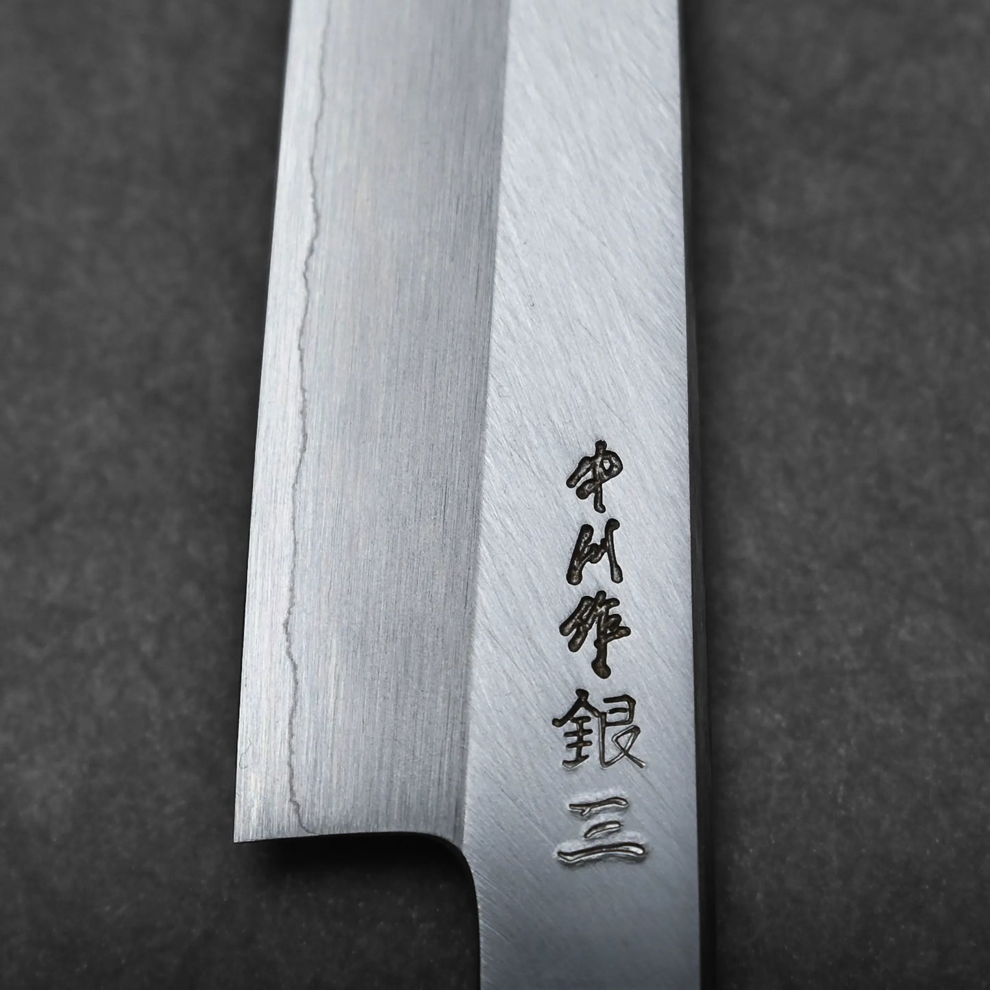 Close-up of a Japanese utility knife blade with a visible wavy pattern and engraved Japanese characters near the edge, set against a dark background.