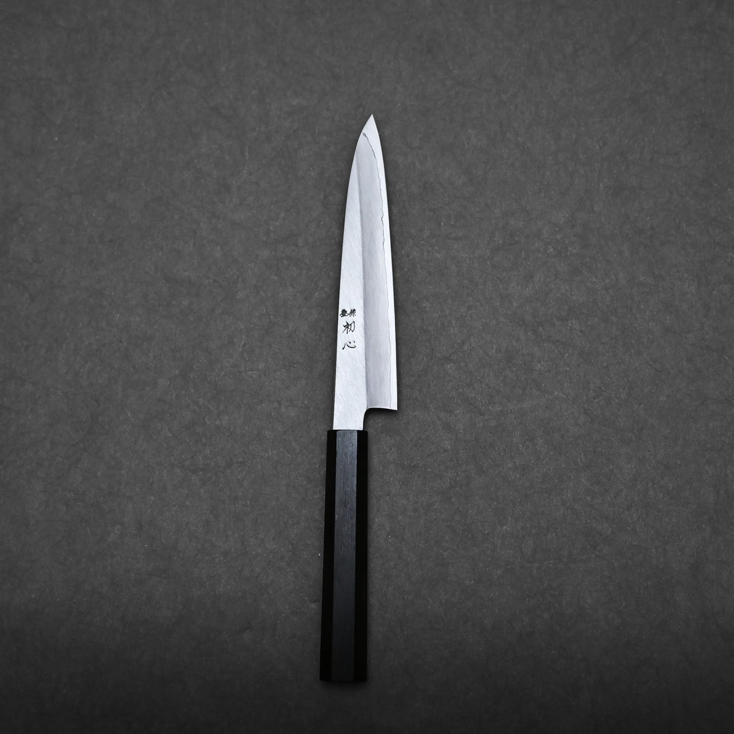 A Japanese utility knife with a sharp, pointed blade and black handle rests on a dark, textured surface. Japanese characters are engraved on the blade of this elegant petty knife.