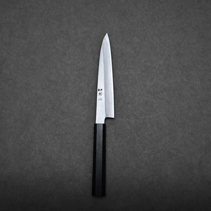 A Japanese utility knife with a sharp, pointed blade and black handle rests on a dark, textured surface. Japanese characters are engraved on the blade of this elegant petty knife.