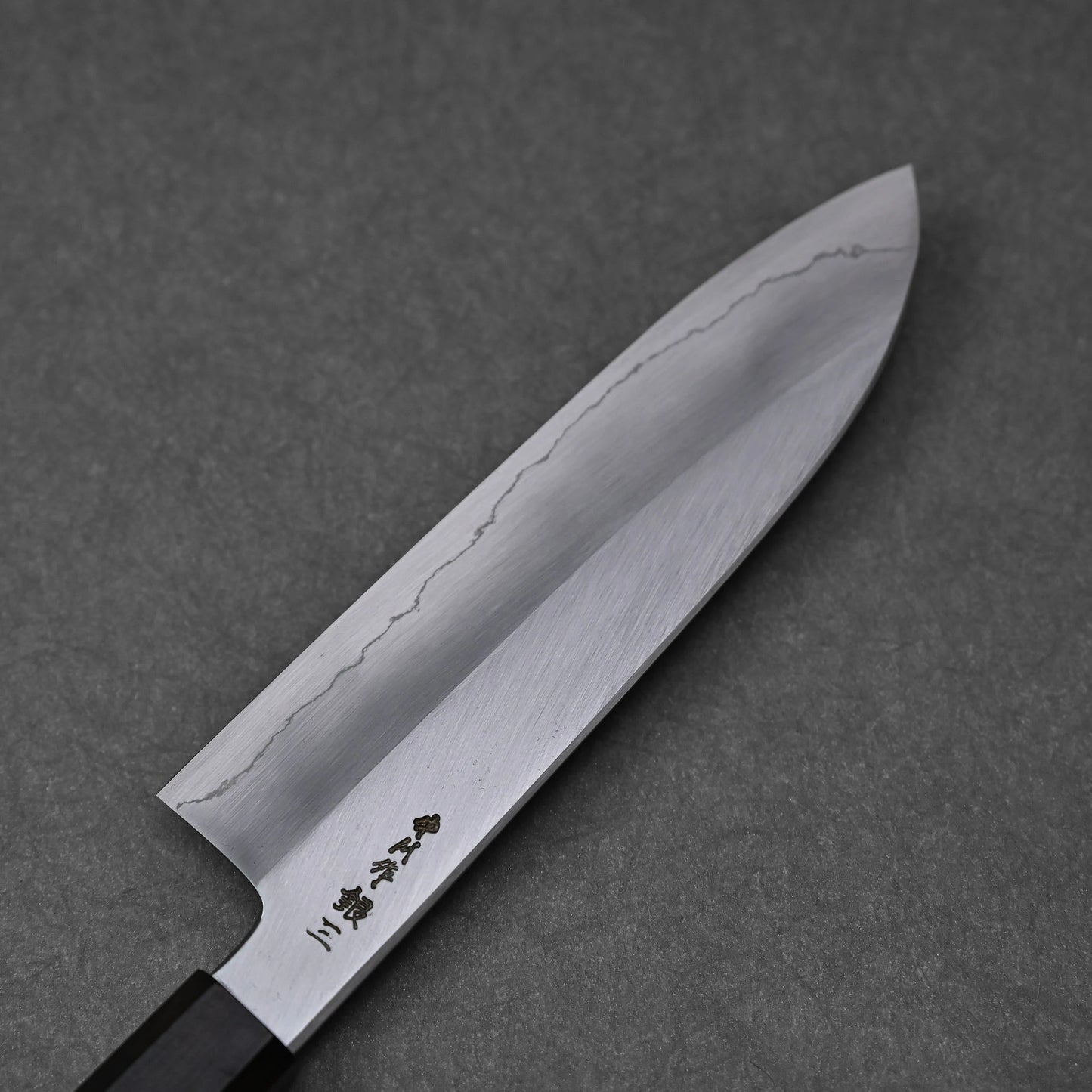 Close-up of the reverse side of a santoku knife blade, showing vertically engraved kanji characters near the handle; photographed to emphasize engraving detail and steel finish.