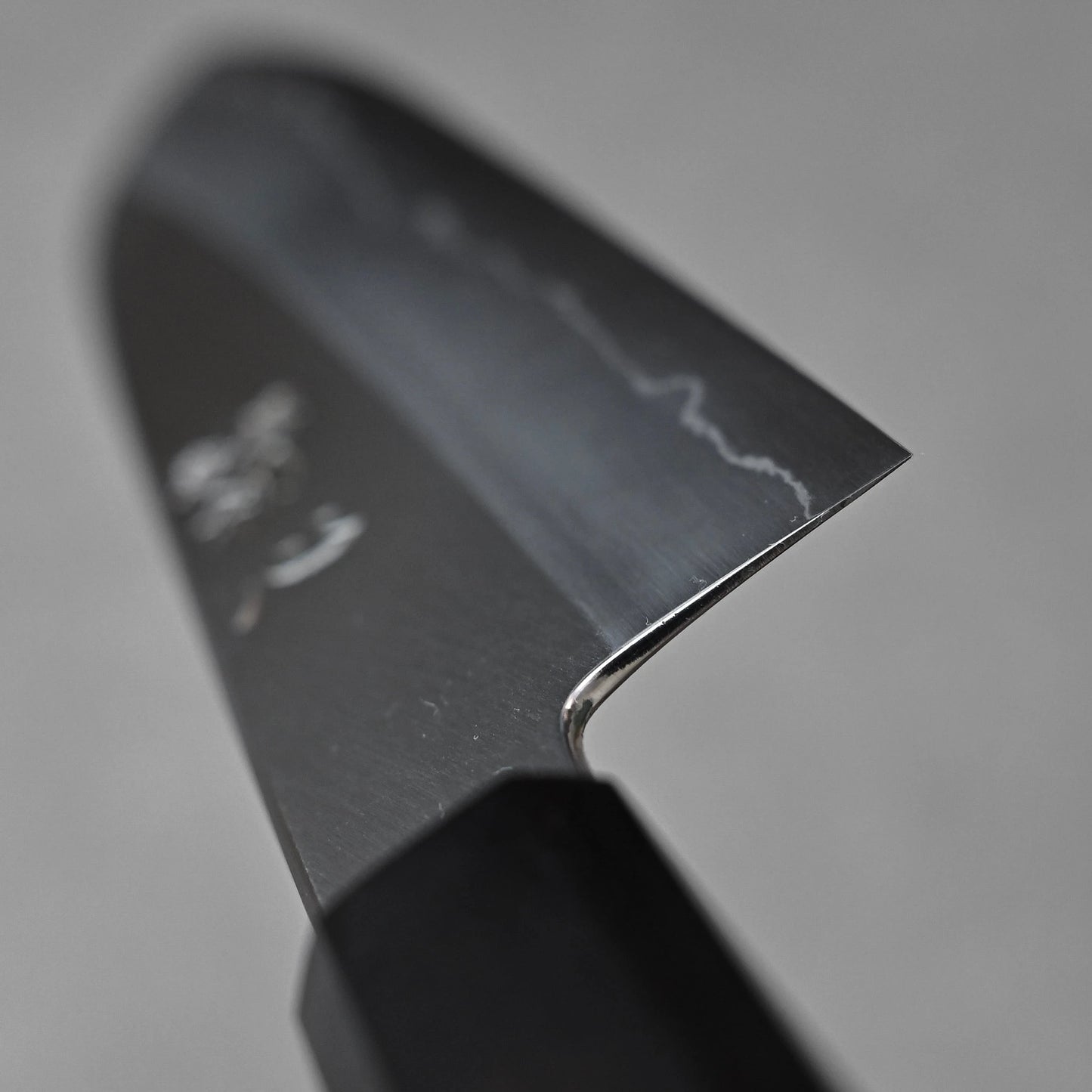 Close-up of a santoku knife blade showing the choil finish near the handle; photographed to emphasize the grind transition, steel surface, and craftsmanship detail.