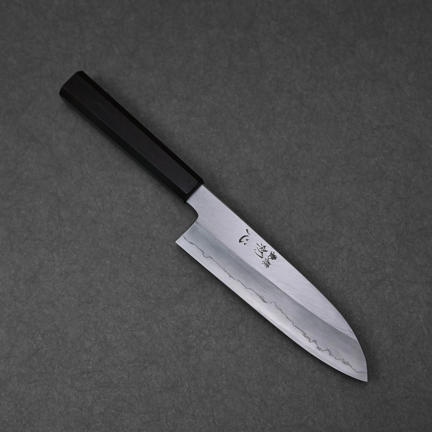 Close-up of a santoku knife blade featuring vertically engraved kanji characters near the handle; photographed to highlight the wavy edge pattern and steel surface detail.