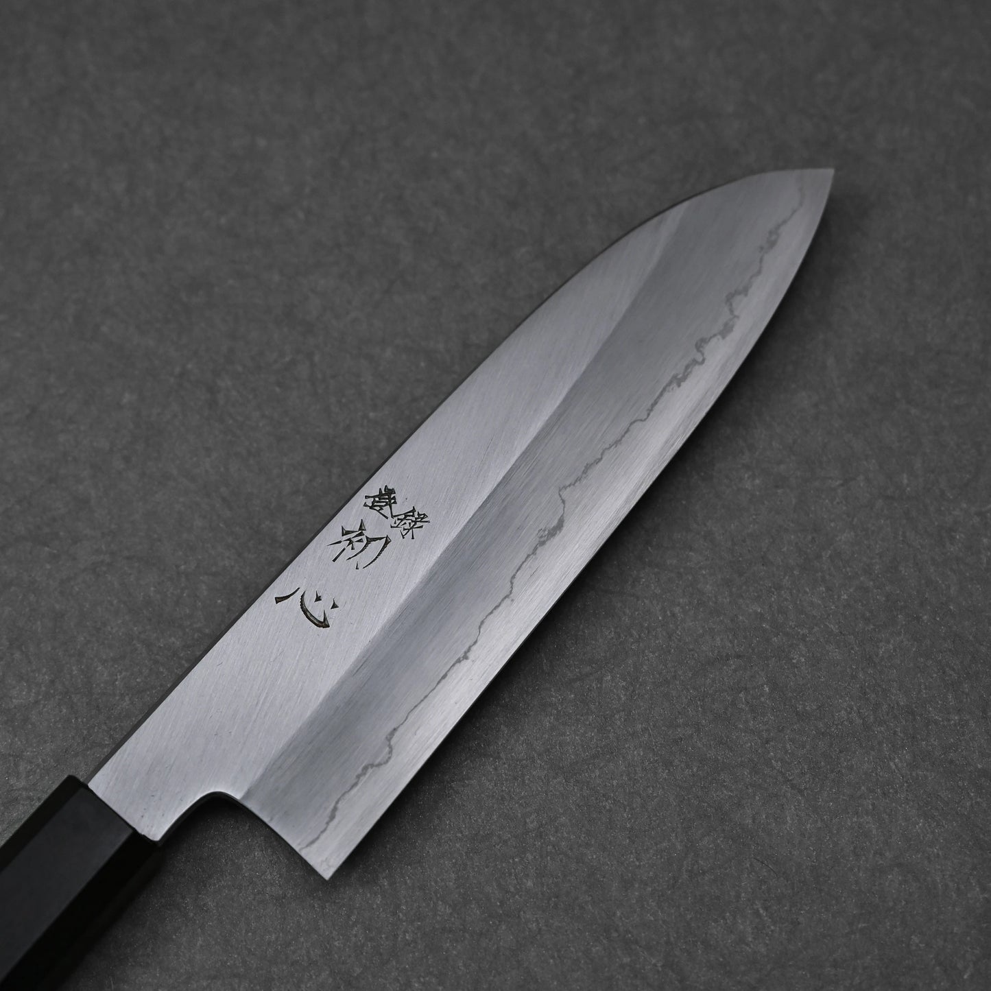 Santoku knife featuring a patterned blade with vertically engraved kanji characters near the handle; photographed close-up to highlight the steel texture and engraving detail.