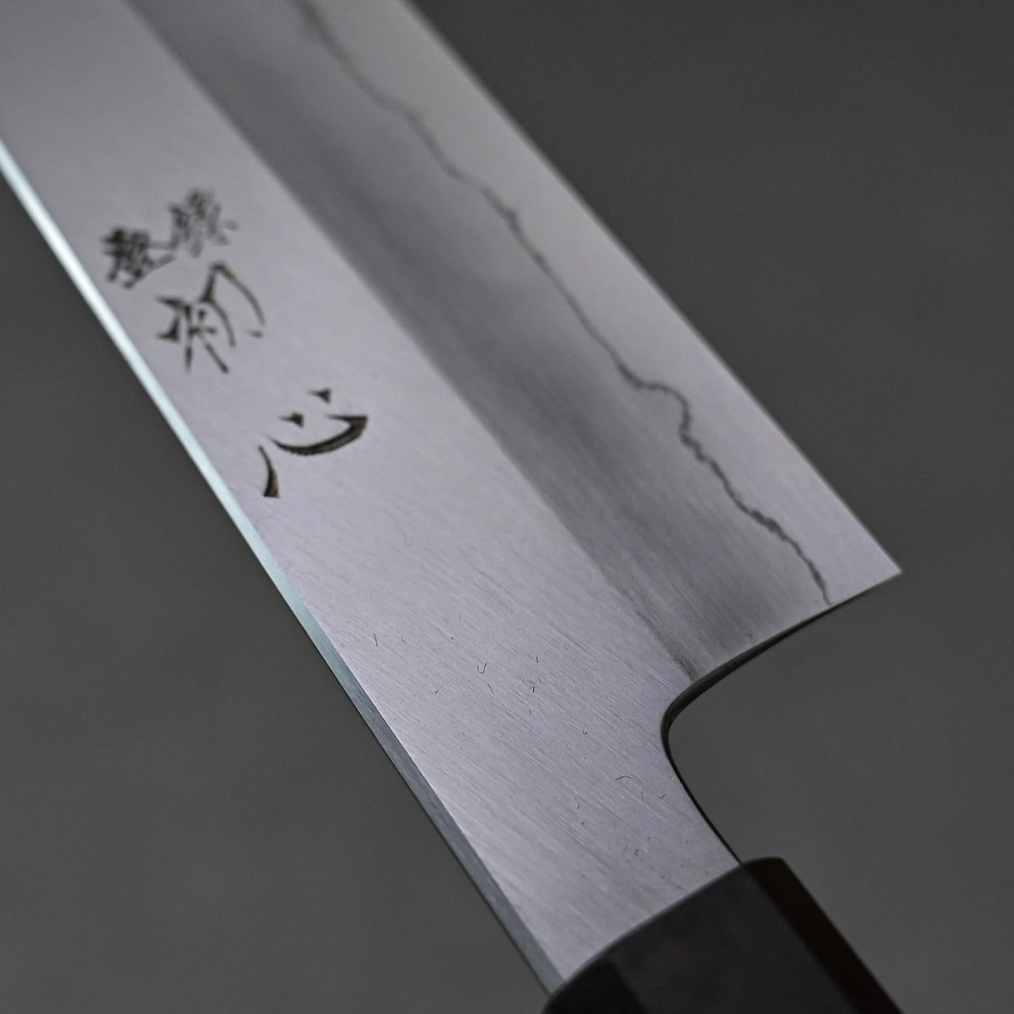 Close-up of a santoku knife blade featuring vertically engraved kanji characters near the handle; photographed to emphasize steel texture and engraving detail.