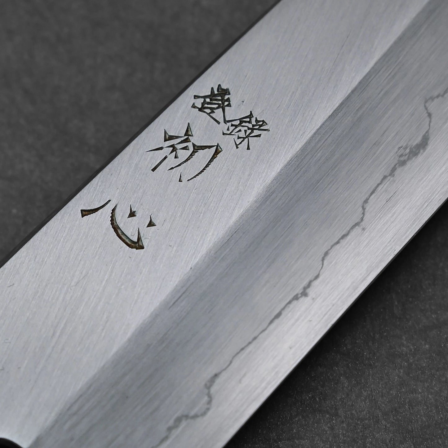 Close-up of vertically engraved kanji characters on a santoku knife blade; photographed to emphasize engraving detail and steel surface.