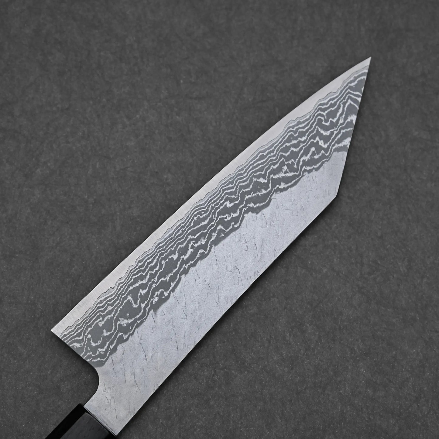 A close-up of the Nigara tsuchime damascus VG10 bunka 180mm chef’s knife blade, showcasing its intricate damascus pattern and wavy lines on VG10 steel, set against a dark gray textured background.