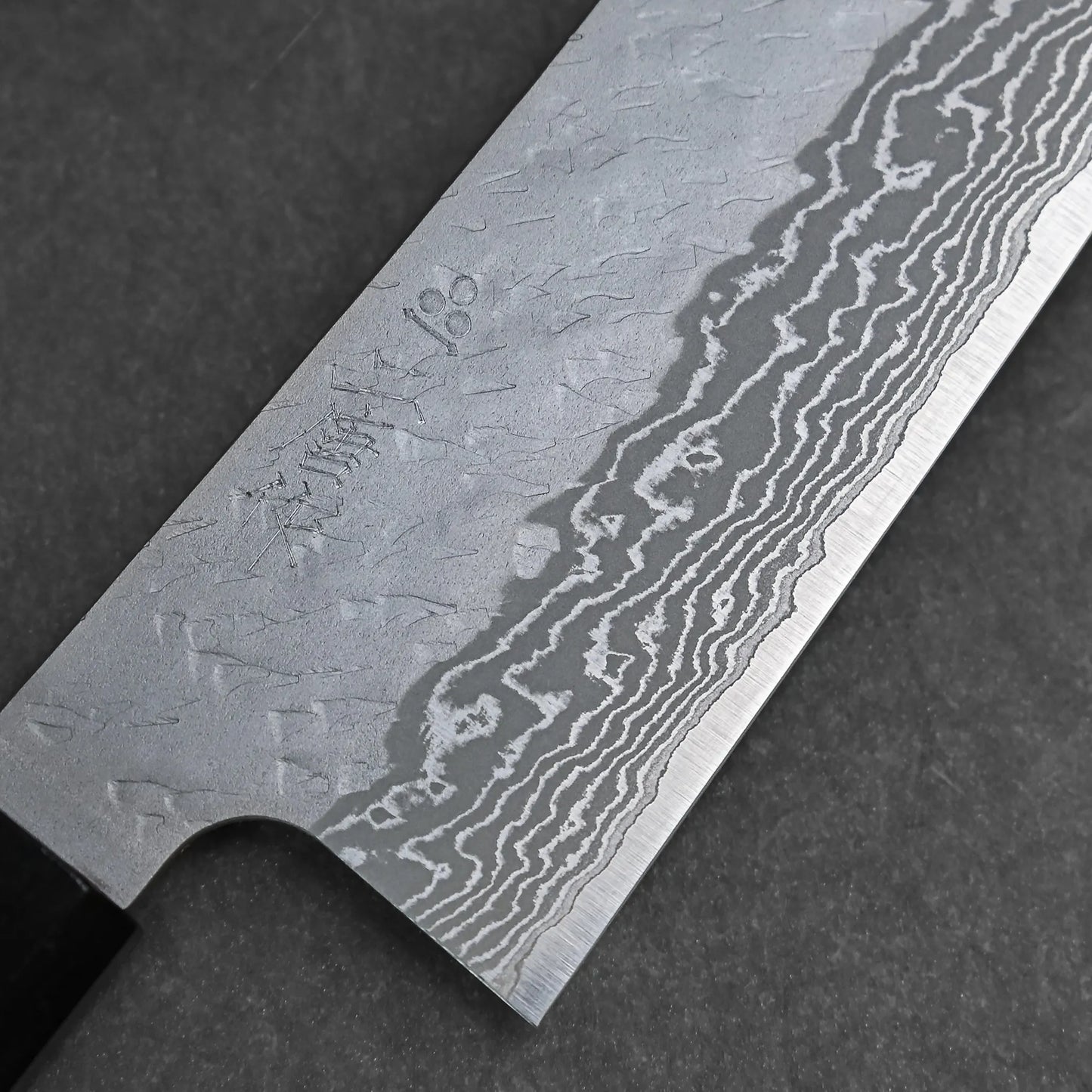 Close-up of a Nigara tsuchime damascus VG10 bunka 180mm knife, showcasing its hammered damascus blade with engraved markings near the handle, displayed on a dark, textured surface.