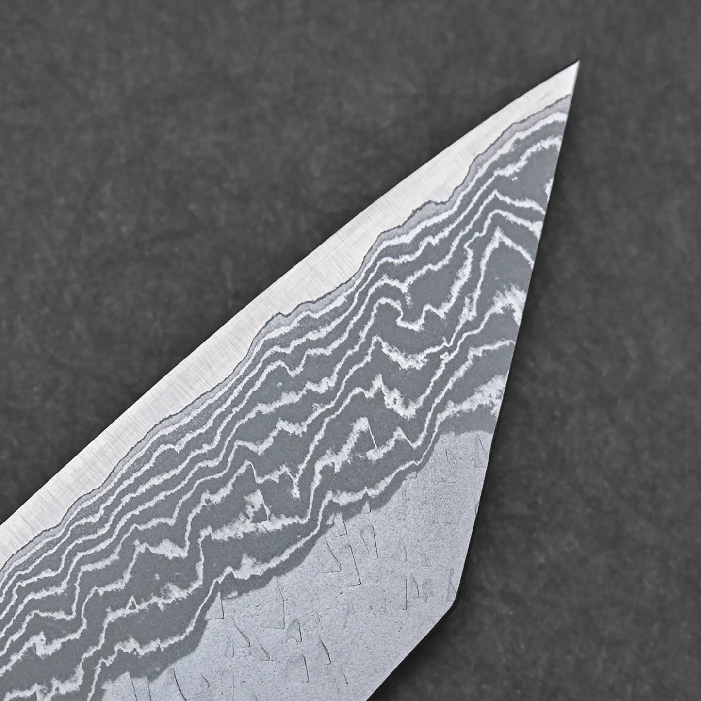Close-up of the pointed tip of the Nigara tsuchime damascus VG10 bunka 180mm, showcasing its wavy patterns. The dark, textured background emphasizes craftsmanship inspired by Nigara Hamono traditions.