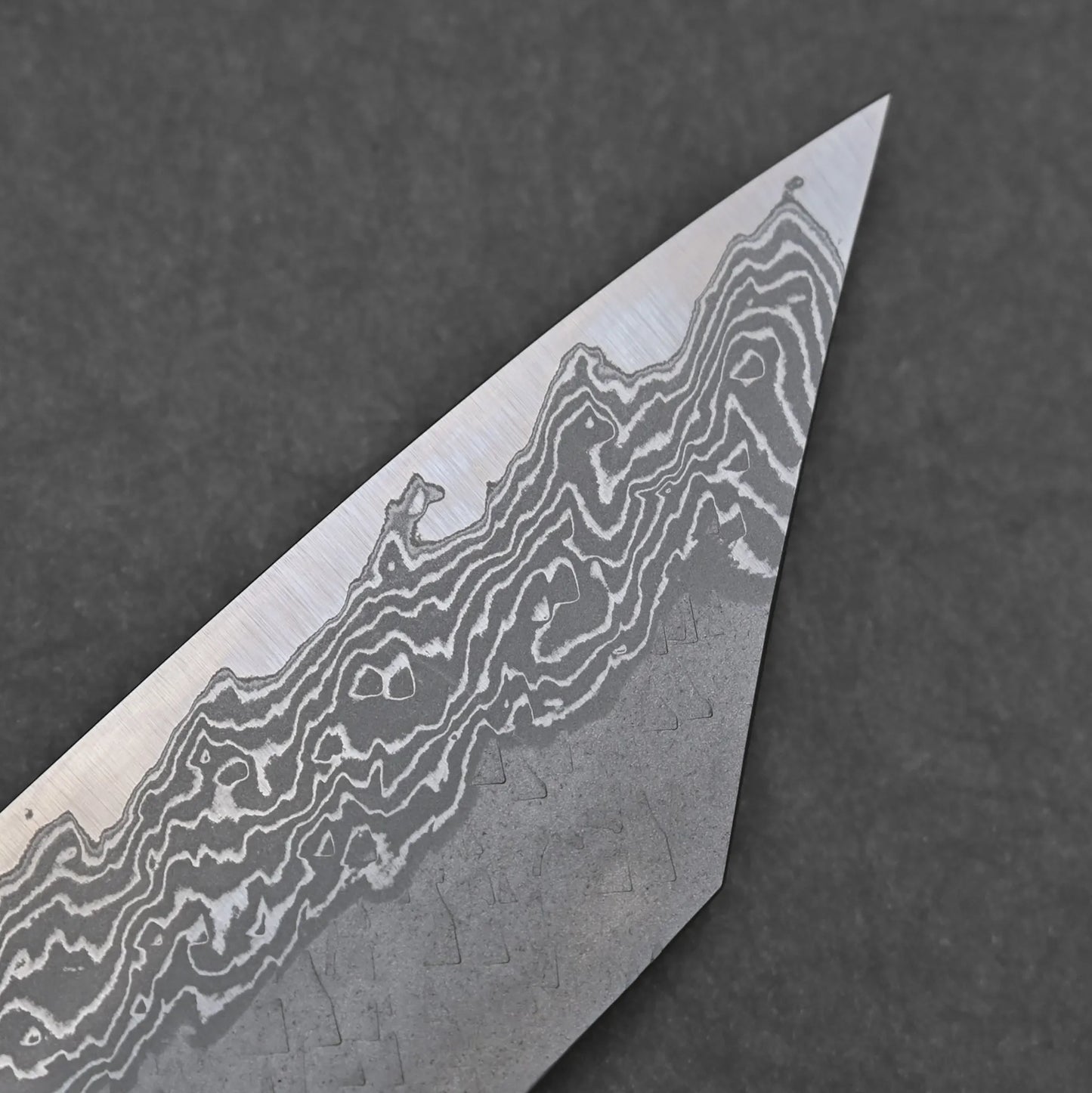 Close-up of the Nigara Tsuchime Damascus VG10 Kiritsuke Gyuto 210mms pointed blade tip, highlighting its unique wavy damascus patterns against a dark gray background.