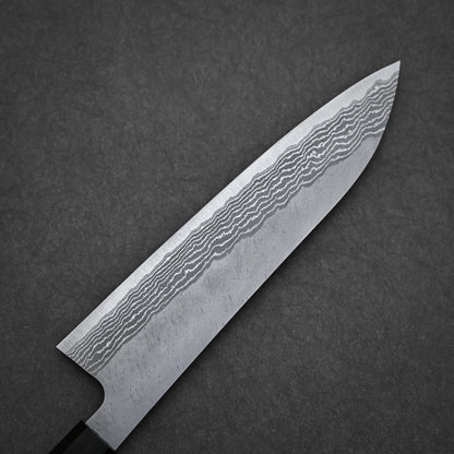 A close-up of the Nigara Hamono Nigara tsuchime damascus VG10 gyuto 210mm, showcasing its wavy Damascus steel blade pattern, rests on a dark textured surface.