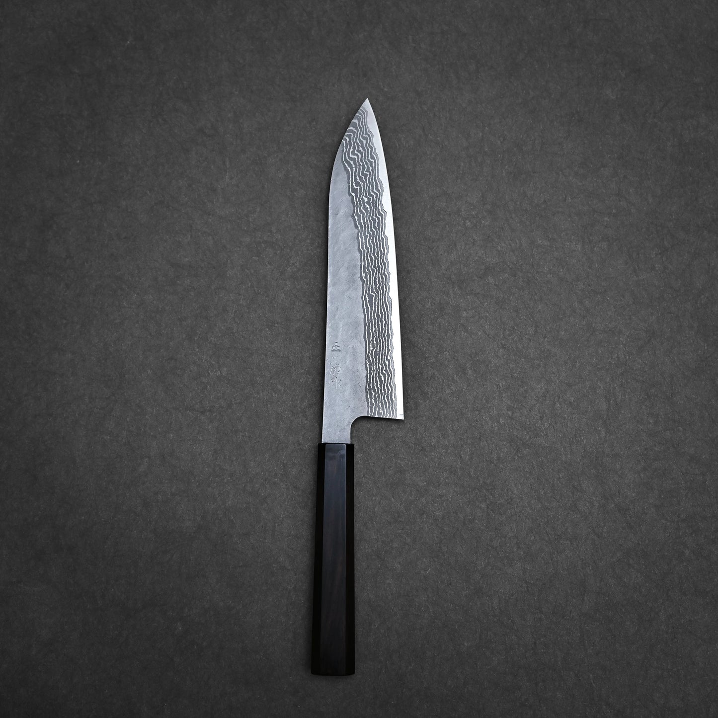A Nigara Hamono Nigara tsuchime damascus VG10 gyuto 210mm knife with a dark handle rests on a plain, dark gray textured surface.