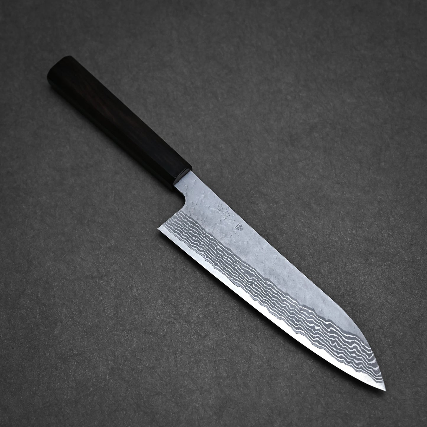 The Nigara Hamono Nigara tsuchime damascus VG10 gyuto 210mm, featuring a dark handle and a wavy-patterned blade, rests on a dark textured surface.