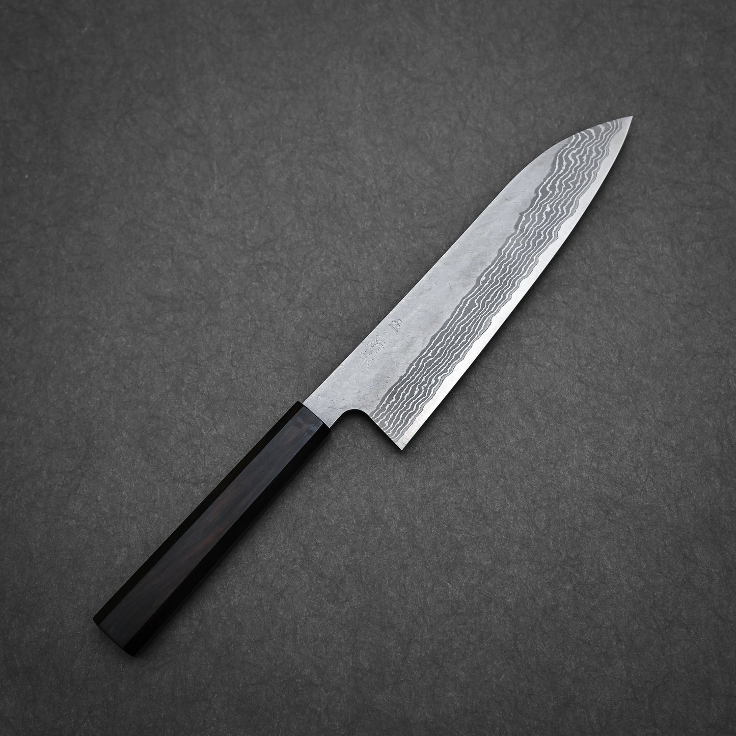 The Nigara Hamono Nigara tsuchime damascus VG10 gyuto 210mm knife, featuring a dark wooden handle and tsuchime-patterned steel blade, rests on a dark gray textured surface.